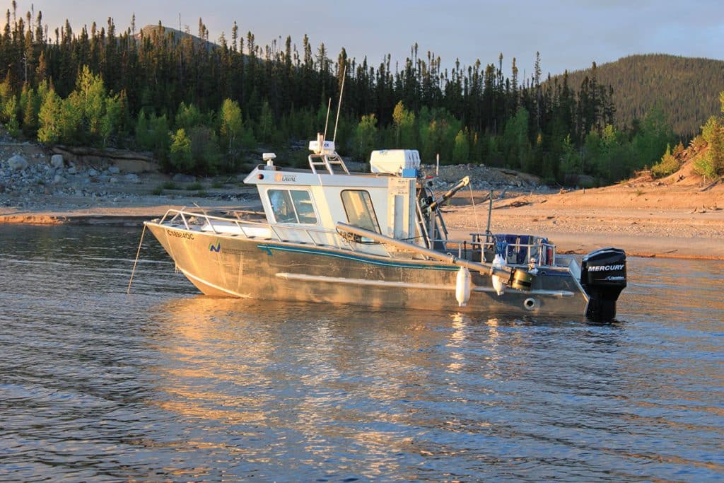 Un petit bateau à moteur, doté d'une cabine de pilotage, vu de profil. Il est ancré près d'une plage de sable blond. Derrière la plage, des conifères et une colline.