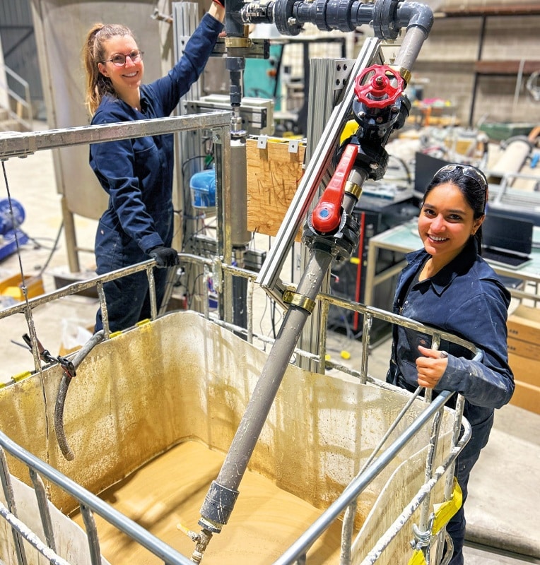 Dans un atelier industriel, deux femmes habillées en combinaison bleue. Elles sourient à la caméra. Elles sont autour d'un réservoir de plastique de forme cubique assez volumineux, posé sur le sol et rempli d'une boue de couleur beige. Le réservoir est renforcé par une cage faite de tubes de métal. Un tuyau est plongé dans le bac. Sur le tuyau, il y a différentes poignées rouges.