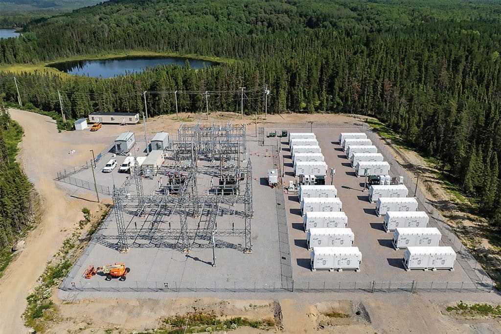 Photo: Vue en plongée d'un poste électrique. La moitié de l'espace est occupée par des conteneurs blancs posés au sol. Autour, une forêt de conifères. En arrière-plan, un petit lac.