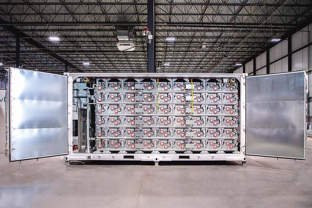 Un conteneur de batteries photographié à l'intérieur d'un bâtiment industriel. Les portes du conteneur sont ouvertes. Le châssis de métal argenté renferme plusieurs tiroirs de batteries empilés les uns au-dessus des autres. Les batteries sont connectées les unes aux autres par différents câbles.