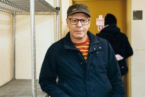 Un homme d'âge moyen se tient debout à côté d'une étagère de métal dont les tablettes sont vides. L'expression de son visage est neutre. Il porte une casquette et des lunettes.