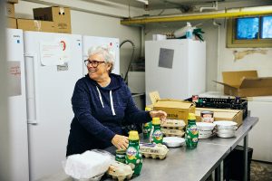 Une pièce dans un sous-sol. Une femme aux cheveux blancs, souriante, se tient debout près d'une grande table. Devant elle se trouvent des boîtes d’œufs et des portions d'aliments préparés emballées individuellement. Derrière elle, plusieurs réfrigérateurs et congélateurs blancs. Des boîtes de carton sont posées sur les réfrigérateurs.