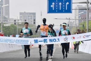 Le fil d'arrivée d'un marathon, dans une grande rue. Quelques coureurs sont devancés par un robot humanoïde. À l'arrière-plan, des panneaux de signalisation routière et quelques bâtiments à plusieurs étages.