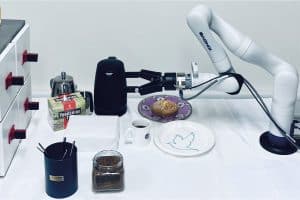 Photo. Un bras robotisé vissé à une table sur laquelle sont posés divers objets: bouilloire, théière, assiettes, tasse de café, pot contenant des bâtonnets pour brasser le café.