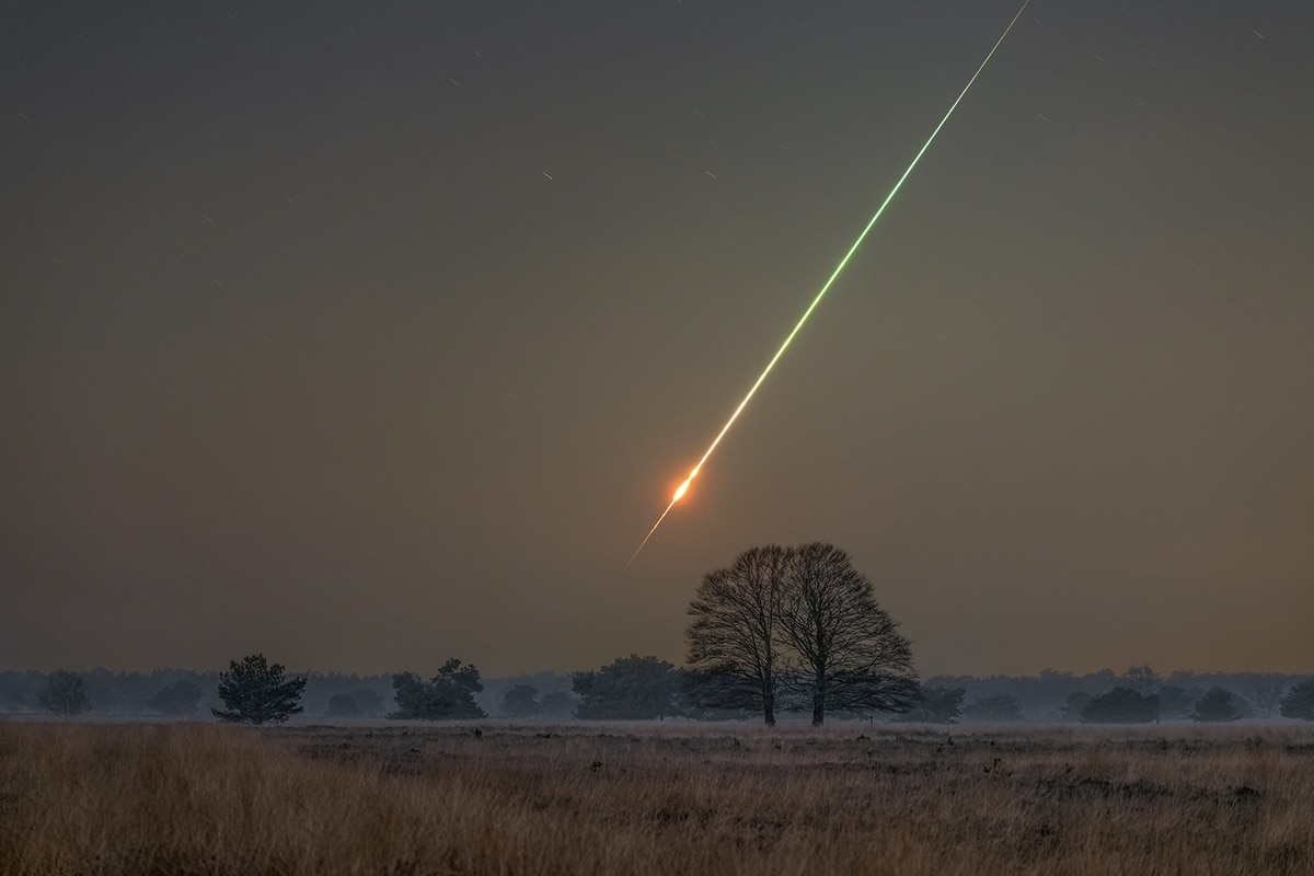 Photo d'un paysage de campagne par temps sombre. Dans un champ, on aperçoit la silhouette de quelques arbres isolés dans un champ. Une traînée lumineuse diagonale traverse l'image du coin supérieur droit au centre de l'image. Au centre de l'image, la lumière est orangée et plus intense.