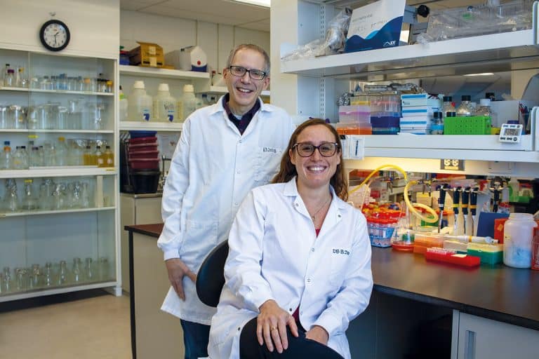 Une femme en sarrau assise dans un laboratoire. Derrière elle, un homme debout, portant aussi un sarrau. Les deux sont souriants.