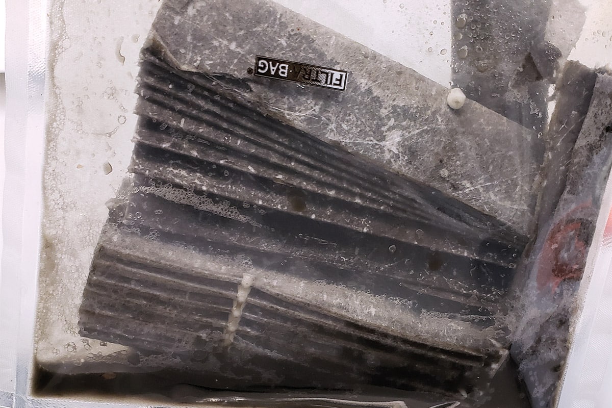 Photo: Un dispositif en forme d'accordéon, gris foncé, portant une étiquette FILTRABAG. Il est inséré dans un sac de plastique transparent. Des gouttelettes d'eau sont visibles sur la surface intérieure du sac de plastique. Au fond du sac, un liquide noir s'est accumulé.