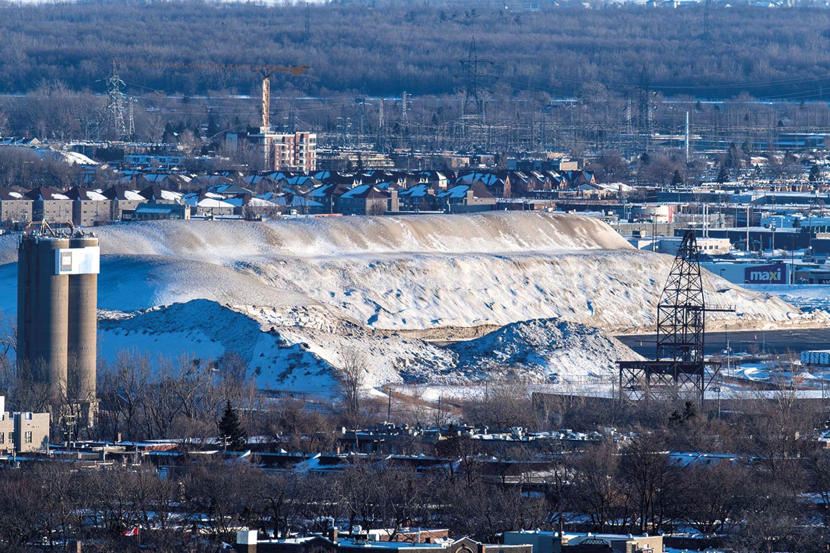 Photo prise par un jour ensoleillé: paysage hivernal en milieu urbain. Une grande montagne de neige est au centre de l'image. Différents bâtiments industriels ou magasins à grande surface sont visibles aux alentours, de même que quelques arbres dépourvus de leur feuilles.