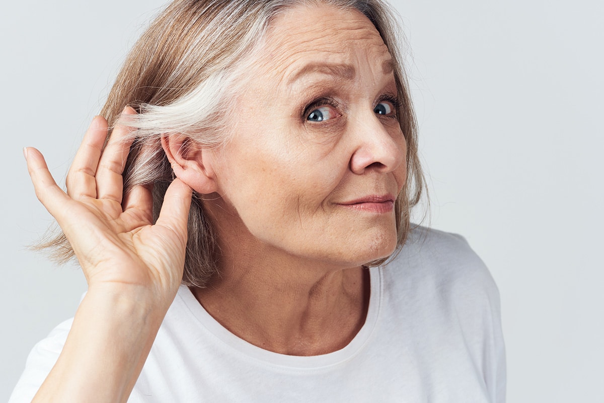 Photo sur fond blanc. Une femme âgée portant un t-shirt blanc tend l'oreille.